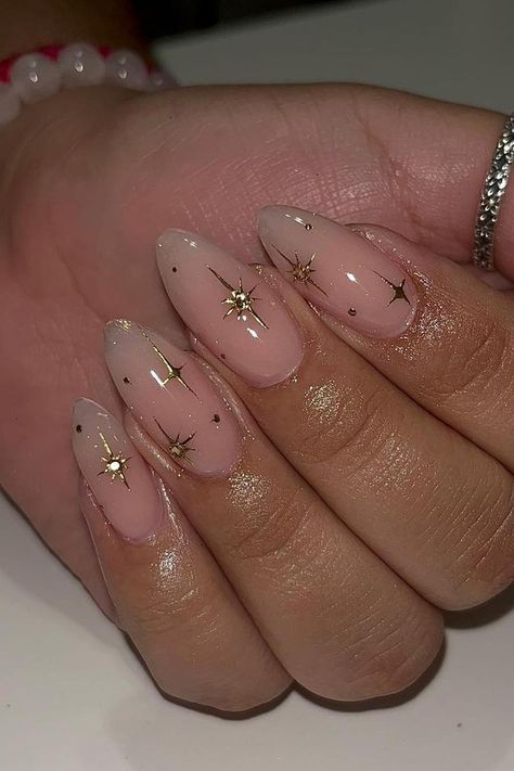 Elegant almond-shaped nails with a translucent pink base, subtly enhanced with tiny gold star accents and specks of gold glitter creating a minimalistic yet chic look. The design is understated and perfect for adding a touch of sparkle to everyday style. Simply charming! ✨  // Photo Credit: Instagram @nailsbykarenm_ Almond Nails Gold Stars, Charm Nails Almond, Almond Shaped Star Nails, Simple Nails Stars, Gold Star Acrylic Nails, Pink Nails With Gold Design, Neutral Nails With Stars, Almond Nails For Graduation, Gold Star Nail Design