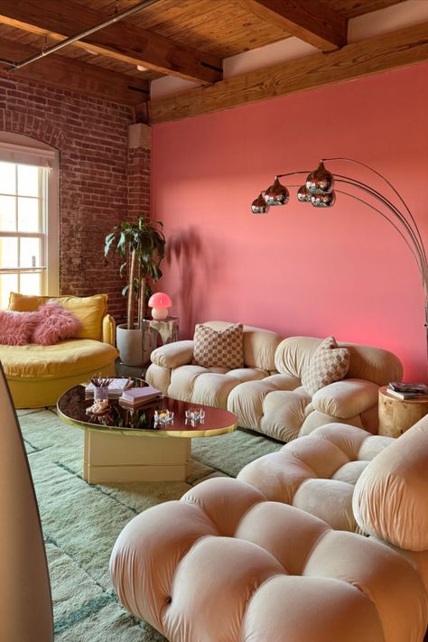 A vibrant living room with a bold pink accent wall, exposed brick, and wooden ceiling beams. It features plush beige tufted sofas, a yellow couch with a pink fluffy pillow, a stylish coffee table with books, and a sleek floor lamp with globe lights. A green plant adds a touch of nature. The room is bathed in natural light from a large window, creating a cozy and chic atmosphere perfect for summer lounging. Patchwork, Colorful Girly Living Room, Hot Pink Couch, Loft Apartment Living Room, Pink Accent Wall, Patchwork Decor, Vibrant Living Room, Girl Apartment, Living Room Transformation