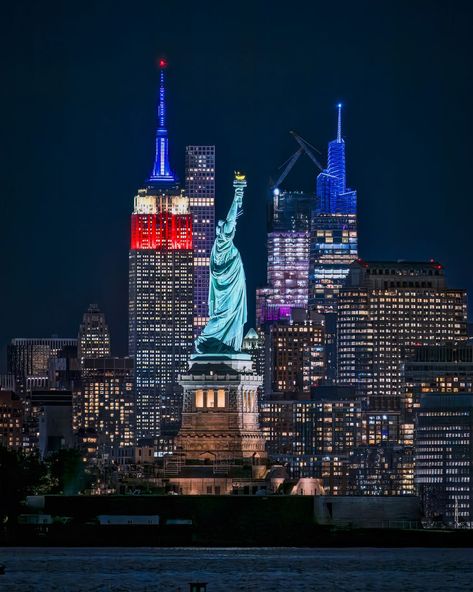 View of the Statue of Liberty in New York Harbor with the Empire State Building lit up in red, white and blue colors Marfa Lights, New York City Pictures, New York Harbor, The Empire State Building, The Statue Of Liberty, City Pictures, Pink Wallpaper Iphone, Solo Female Travel, Blue Colors