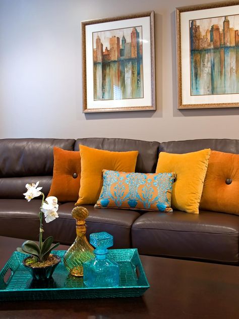 A brown leather sofa matches a dark wooden coffee table in front of the neutral living room walls. Orange throw pillows and a contrasting patterned blue pillow add color to the room. Two scenic photos and a bright blue breakfast tray pull repeat the hue. Brown Sofa Living Room, Turquoise Room, Brown Couch Living Room, Brown Leather Couch, Brown Living Room Decor, Brown Couch, Living Room Orange, Living Room Warm, Brown Leather Sofa