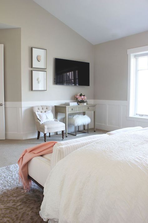 Gorgeous bedroom features beige paint on upper walls and wainscoting on lower walls framing flatscreen TV over curved gray console table next to stacked art above beige tufted chair. A queen bed is dressed in soft white bedding as well as striped sofa placed at the foot of the bed. Decorating Around A Tv, Bedroom Wainscoting, Wall Decor Living Room Modern, Wainscoting Bedroom, Tv Ideas, Wainscoting Styles, Diy Wall Decor For Bedroom, Nursery Frames, Bedroom Tv