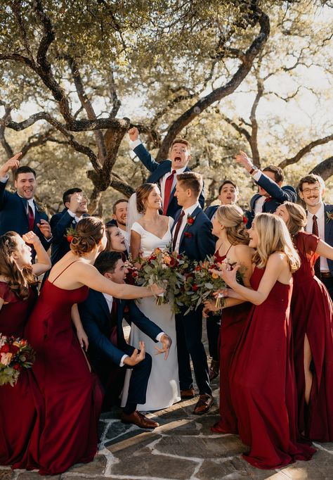 Fun excited wedding party surrounding bride and groom on wedding day wearing red bridesmaid dresses and groomsmen in blue suits Wedding With Bridesmaids And Groomsmen, Winter Wedding Red Bridesmaid Dresses, Theme Ideas For Wedding, Deep Red Groomsmen Attire, Navy Blue And Red Groomsmen Suits, Red Wedding Bridesmaid Dress, Red Bridal Party Color Schemes, Wedding Deep Red, Red And Blue Rustic Wedding
