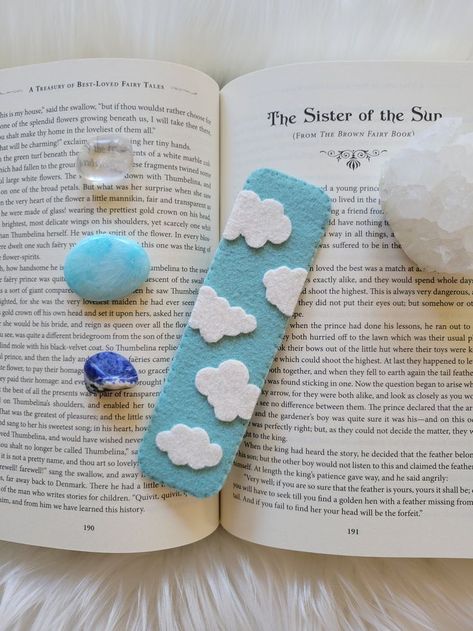 A light blue bookmark with white clouds sitting ontop of a book, surrounded by crystals. Fimo, Foam Sheet Bookmarks, Wool Ideas Crafts, Sewing Bookmarks Diy, Bookmark Clouds, Bookmark Handmade Ideas, Punch Needle Bookmark, Felt Book Marks, Bookmarks Handmade Easy