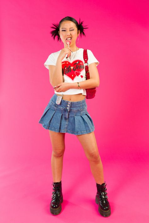 Girl standing in front of a pink backdrop licking a lollipop with her face scrunched up and one eye closed. She is wearing a white with jean top with a red heart on the front, pleated denim skirt, red vintage guess shoulder bag and black platform buckled doc martins. Early 2000s Editorial, Vintage Outfit Photoshoot, Girly Aesthetic Photoshoot, Y2k Fashion Models, Trashy Y2k Photoshoot, Y2k Fashion Editorial, 2000s Music Video Outfits, Y2k Editorial Photoshoot, Fashion Campaign Photography