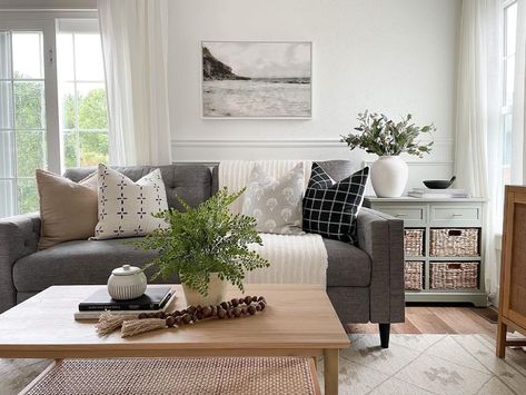 This brightly lit living room showcases a dark grey couch decorated with brown, grey, white, and black throw pillows. A light wood coffee table sits in front of the sofa accessorized with stacked books and a potted plant. A light green side table with woven baskets rests on the side of the sofa. Dark Grey Couch Wood Coffee Table, Grey Couch And Coffee Table, Black And White Throw Pillows Grey Couch, Dark Gray Couch Green Pillows, Dark Grey Couch, Charcoal Couch, Grey Couch Decor, Grey Sectional Couch, Dark Couch