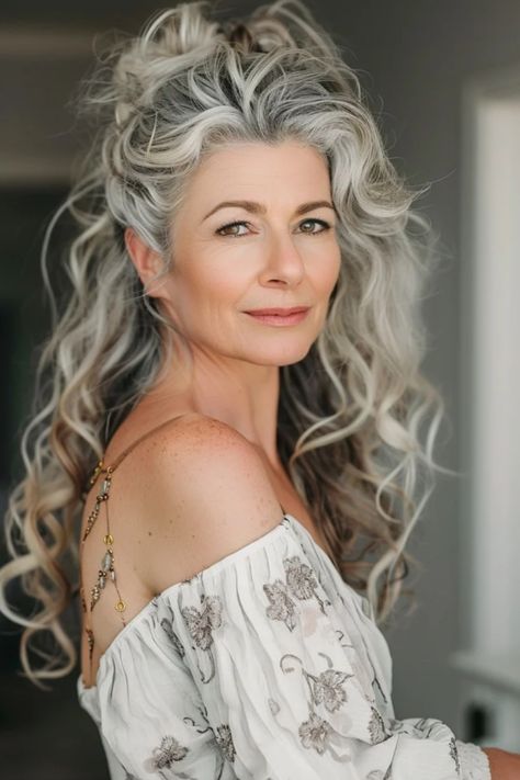 Elegant older woman with long, wavy gray hair and a white floral off-shoulder top, looking over her shoulder with a gentle smile. Short Curly Hair For Older Women Over 50, Gray Hair Mixed With Blonde, Womens Up Do Hairstyles, 2024 Summer Haircuts For Women, Grey Hair Styles For Women Over 50 Long, Over Fifty Hairstyles Over 50, Over 50s Hairstyles, Long Curly Hair Over 50 Women, Grey Curly Hairstyles