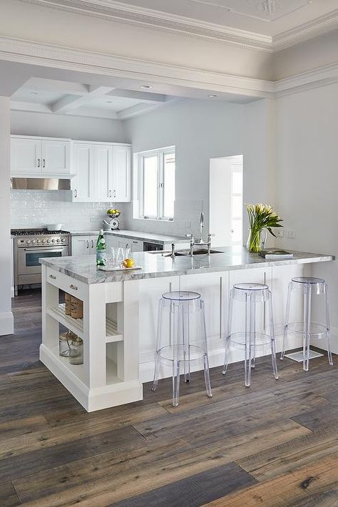 Backless acrylic stools sit in front of a white kitchen peninsula accented with slated shelves and a Super White Dolomite countertop completed with a stainless steel dual sink and a polished nickel gooseneck faucet fixed facing l-shaped Super White Dolomite countertops completed white shaker cabinets located under a window. White Kitchen Peninsula, Kitchen With White Cabinets, Kitchen Peninsula, Smitten Kitchen, Classic Kitchen, White Kitchen Design, Kitchen Design Decor, Kitchen Remodeling Projects, Chic Kitchen