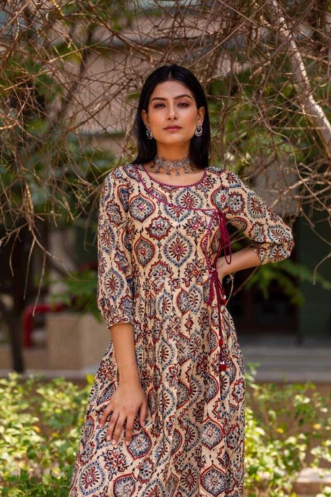 Exemplifying the age old art of Ajrak printing in a medley of light and dark tones, this Kurta is styled in an Angrakha silhouette. Flawlessly embellished with mirror and thread work on the yoke and along neck line, the angrakha is enclosing with a double tie dori that finishes with beautiful cowry and pom-pom tassels. Ajrak Print Kurti Designs, Ajarak Print Kurta Design, Ajarak Print Kurta, Ajrak Kurti Designs, Sleevless Kurti, Angrakha Style Kurti, Angrakha Style Kurta, Business Casuals, Simple Motif