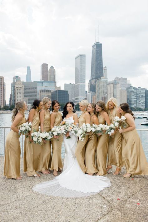 Nine bridesmaids in silky long gold dresses stand on either side of the bride, looking at her. Everyone is holding a bouquet made by Romee Willow Florals. The Chicago skyline is in the background. Vintage Gold Bridesmaid Dresses, Yellow And Gold Bridesmaid Dresses, Cream White Bridesmaid Dresses, White And Gold Bridesmaid Dress, Gold And White Wedding Bouquet, White And Gold Wedding Theme Bridesmaid, Gold Yellow Bridesmaid Dresses, White Green Gold Wedding Bouquet, Yellow Gold Bridesmaid Dresses
