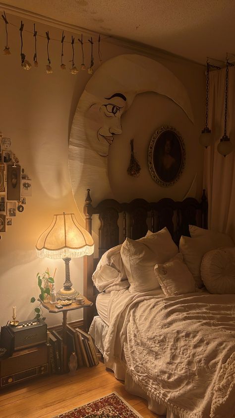 A cozy bedroom with an antique wooden bedframe topped with a vintage cream blanket and a cozy mix of pillows. Above the bed hangs a paper moon and dried flowers as well as old swag lights. To the left of the bed is a pretty vintage lamp on a small table filled with knickknacks and candles. Below, lent against the side are books and an old radio. Unique Room Layout Bedroom, Canopy Bed With Wallpaper, Art Next To Bed, Feminine Bohemian Decor, Stevie Nicks Bedroom, Whimsigoth Small Bedroom, Bedroom Ideas Whimsigoth, Minimalist Whimsical Bedroom, Earthy Bedroom With Carpet