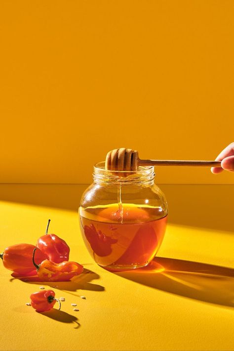 hot honey dripping into honey jar with peppers near by on yellow backdrop