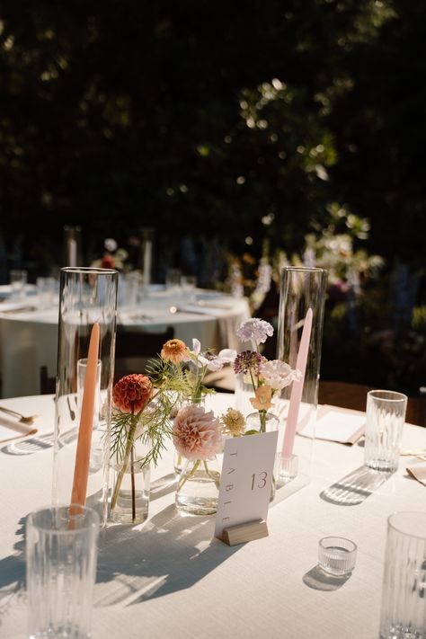Summer garden party wedding in PNW with free form and vibrant florals. Center pieces with bud vases and a variety lf stems. Hurricanes and multi colored taper candles staggered in centers. Flowers On Round Table, Bud Vases For Wedding Tables, Wedding Simple Flower Centerpieces, Flower Buds Centerpiece, Colored Tapered Candles, Garden Wedding Centerpieces Long Table, Centerpieces Wedding Not Flowers, Round Table Taper Candles Wedding, Diy Wedding Bud Vases