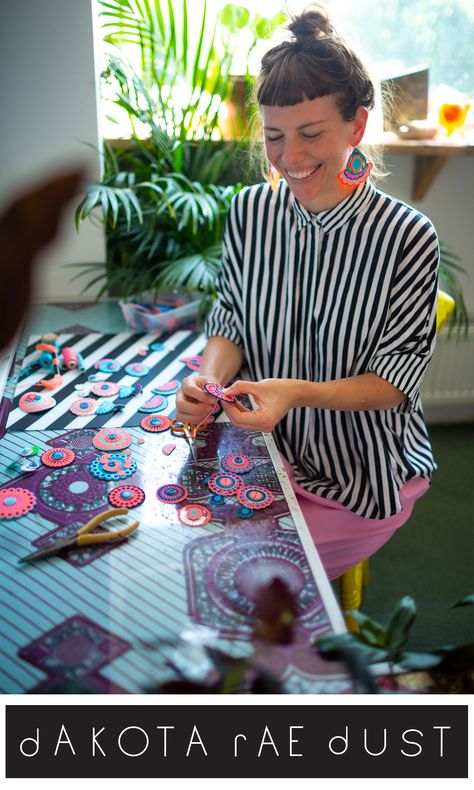 Jewellery designer Bec is sitting at a desk scattered with colourful patterned discs. The desk has a blue and purple pattern on it and a pair of pliers are seen next to the discs. She is wearing a black and white striped shirt and large, colourful teardrop shaped earrings. She is looking down focusing on the circular patterned earring component she is hand stitching and smiling. In the background a large house plant is visible. Textile Fiber Art Jewelry, Fabric Bead Earrings, Earrings From Fabric, Textile Jewellery Handmade, Scrap Fabric Earrings, Textile Necklace Wearable Art, Textile Jewelry Diy, Jewelry Making Studio, Fabric Jewelry Necklace