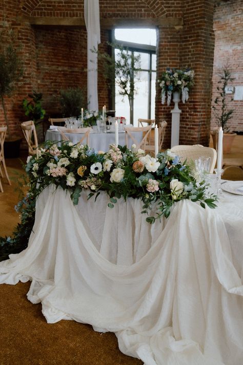 This is an image showing a wedding top table in a barn. On the table are a luxurious layers of drapes and a large floral arrangement. On the table are also glass candlesticks with white candles, tableware and table signage. Behind the table are french style chairs. Fabric Draping Wedding, Table Drapes, Ceiling Drapes, French Style Chairs, Classic Vibes, Draping Wedding, Barn Photography, Fabric Draping, Desert Table