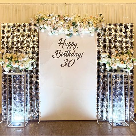 Impart a truly majestic allure into your 30th birthday party by setting up a breathtaking backdrop. Arrange rectangular frames covered in blush spandex cover along with silver sequins wall panels and embellish them further with a lush pastel hued flower arrangement to create a mystically magical effect. Place silver column stands with silver flower pots flaunting a pretty bunch of pastel hued rose, peony, & daisy flowers and create fanciful lighting effect by setting up white backdrop uplights. Women Birthday Backdrop Ideas, Shimmer Wall Backdrop With Flowers, 70 Birthday Decoration Ideas, Birthday Backdrop Ideas For Women, White And Silver Backdrop, Silver Shimmer Wall Backdrop, Bday Backdrop, Sequins Wall, 25th Wedding Anniversary Decorations