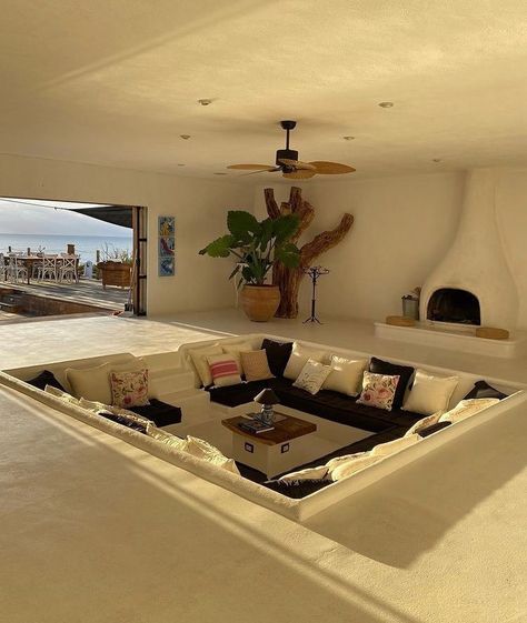 An off-white/cream living room. A set in conversation pit is in the middle of floor with black cushions and around 2 dozen pillows. A fireplace is on the right hand side. A wooden ceiling fan is present. A large plant and tree trunk sit in the top corner. A large opening to the patio is on the left hand side where fold back glass doors are folded back. Dream Rooms, Villa Home, Dream House Rooms, Hus Inspiration, Dream Apartment, Dream House Interior, Dream House Decor, House Inspo, My New Room