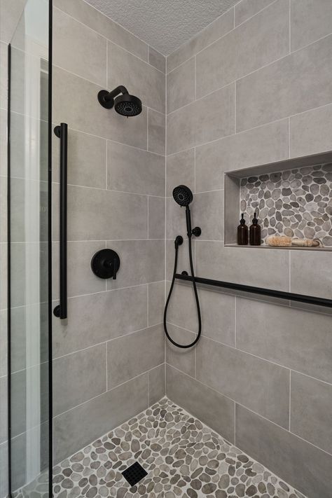 A transitional shower design with river rock floor and large format grey tile on the walls. Matte black fixtures. Large Grey Tiles Bathroom, Modern Farmhouse Tiled Shower Ideas, Tiny Bathroom Renovations, Restroom Tile Ideas, Glass Tile Shower Walls, Subway Tile Showers With Accent, Small Master Shower Ideas, Black And White Shower Ideas, Shower With River Rock Floor
