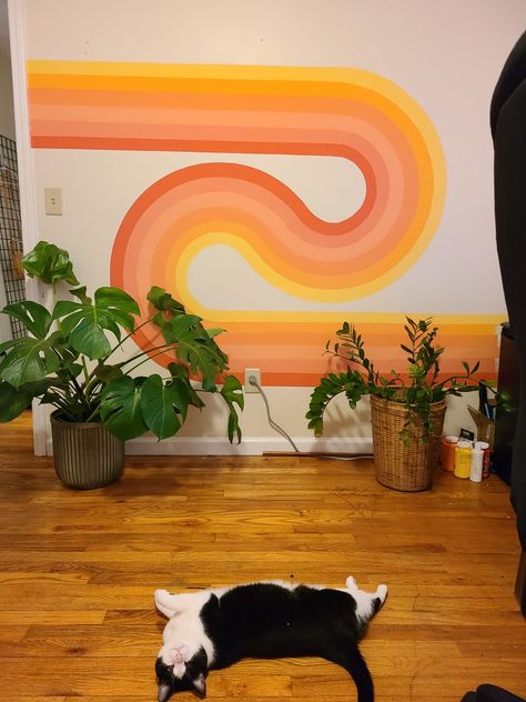 70s wavy wall art with pink peach yellow orange tones in a rainbow looping together with a big monstera plant and zz plant on the right wooden floor with a very cute black and white Cat laying on the floor looking at the camera Mural Wall Art Rainbow, Aesthetic Walls Painted, 70s Feature Wall, Painting Ideas On Wall Aesthetic, Peach And Yellow Aesthetic, 70s Inspired Wall Paint, 70s Wavy Wall Art, Aesthetic Wall Paintings Bedroom, Wall Mural Design Ideas