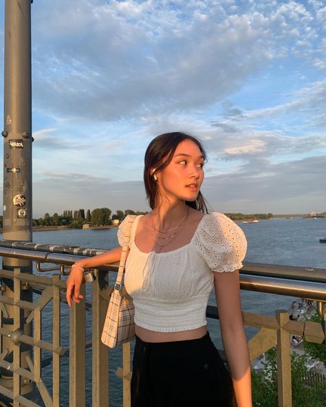 Girl with white puff sleve top and black long pants standing on a bridge. Golden necklace around her neck and a pink bracelet around her hand. She is looking to her right with one arm resting on the bridge railing. A slight smile on her face and sunlight hitting her face. Around her is the beautiful sky and a sea. She is standing on a bridge. There is a see behind her. There is a blue sky with some clouds. Alanya, Pose Ideas On Bridge, Poses For Post Instagram, Picture On Bridge, Leaning Poses Photography, Poses For Photoshoot Models Standing, Poses For Bridge, Pose Reference Photo Instagram, Poses On Bridge Photo Ideas