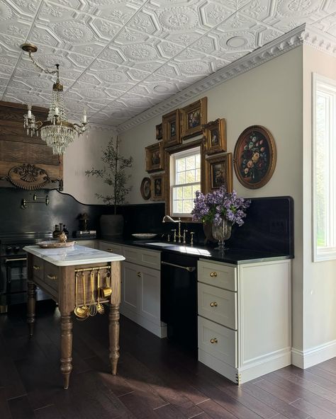 Our Modern Victorian kitchen stills📸 I hope y’all can appreciate the details in the space…the @americantinceilings tiles, the Nero Mist granite counters and tall backsplash with an ogee edge from @graniteworksstone , the antique Italian chandelier, the gorgeous custom dairy table island made by @clevelandcarpentry , the antique floral oil paintings I’ve been collecting , the 1800’s bed frame turned hood vent , the glass bifold pantry with brass @signaturehw Cremone bolts—it all came together ... Brass Hardware Kitchen, Bifold Pantry, Bridgerton Love, Dairy Table, Modern Victorian Kitchen, Modern Victorian Design, Modern Victorian Decor, Modern Victorian Interiors, Modern Vintage Kitchen