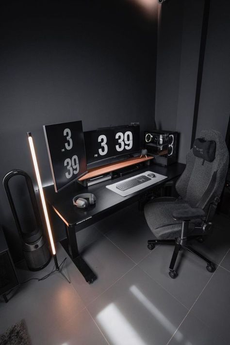 Modern office setup featuring a black desk with dual monitors displaying large digital clocks. The room has a dark, monochrome color scheme, highlighted by vertical ambient light strips that add a warm glow to the space. The setup includes a high-end gaming chair, a laptop, and a desktop computer, all set against a dark grey wall. This setup is designed for both aesthetic appeal and functionality, with a minimalist yet stylish approach. Moody Desk Setup, Minimalist Desks, Dark Desk, Computer Desk Organization, Minimal Desk Setup, Minimal Workspace, Minimal Desk, Positive Environment, Desk Setups