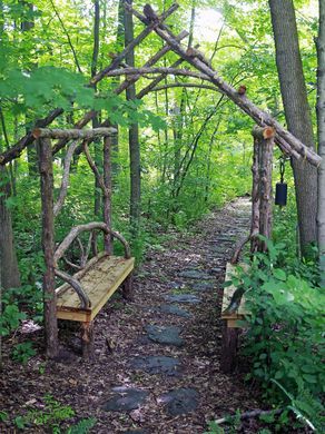 An arbor made of cedar wood with a seating area leads Things To Do With Cedar Wood, Wooded Pathway Ideas, Seating Area In Woods, Paths In The Woods, Secret Forest Garden, Heavily Wooded Backyard Landscape, Wood Trails Pathways, Wood Trails Ideas, Woodland Path Ideas