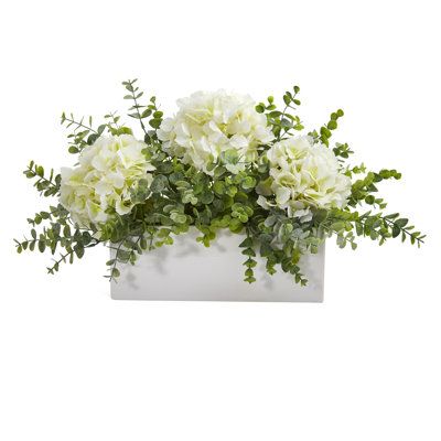 Incorporate hints of countryside beauty with the help of this artificial arrangement flaunting a collection of realistic looking silk hydrangea blossoms, each intricately designed in soft, subtle colors and highlighted throughout with an overgrowth of eucalyptus foliage in complementary green hues. Resting 15in. wide in a white vase, this incredibly lifelike arrangement evokes elegant beauty wherever displayed. About Gracie Oaks Inc. - For over 75 years, Gracie Oaks Inc. has been providing consc Advent Church Decorations, Hydrangea And Eucalyptus, White Hydrangea Centerpieces, Hydrangea Flower Arrangements, Rectangular Vase, Spring Flower Arrangements, Hydrangea Centerpiece, Hydrangea Arrangements, Unique Flower Arrangements