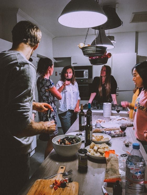 a group of people standing around a kitchen table with food on the counter and wine bottles