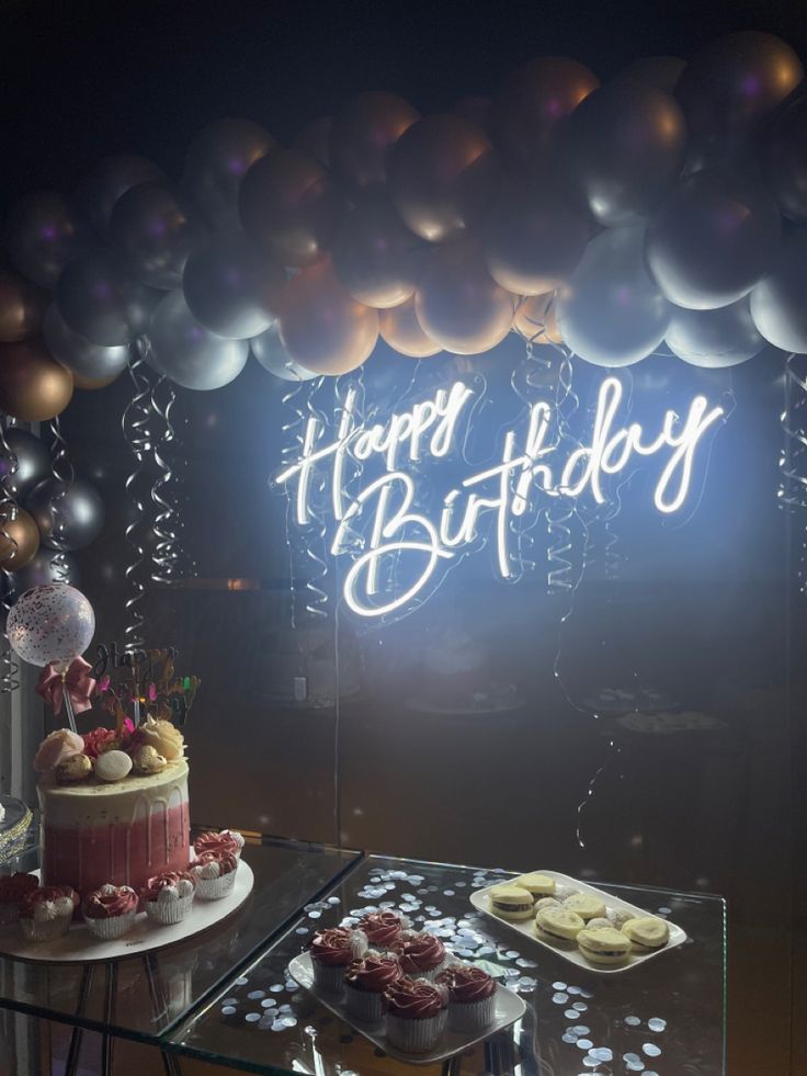 a birthday cake and cupcakes sit on a table in front of a neon sign that says happy birthday