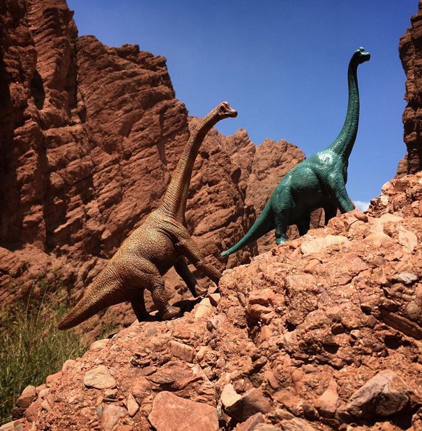 two toy dinosaurs are standing on rocks in front of some mountains and boulders, one is green and the other is brown