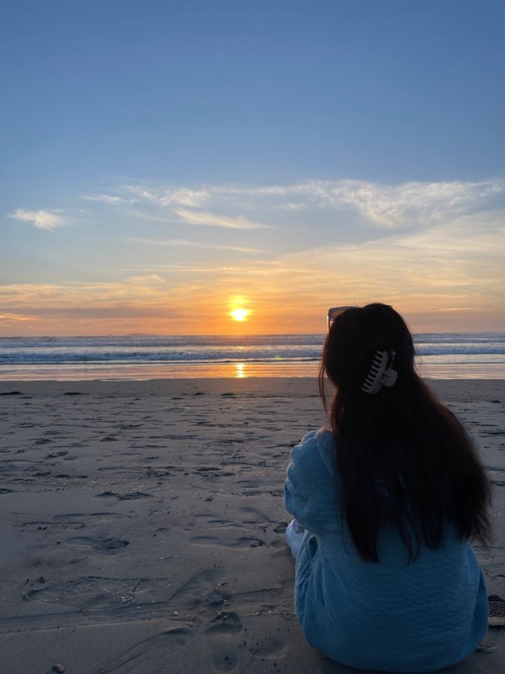 a woman sitting on top of a sandy beach next to the ocean under a sunset