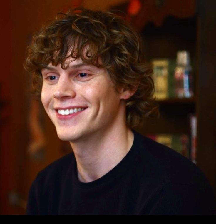 a smiling young man with curly hair and blue eyes looks at the camera while wearing a black t - shirt