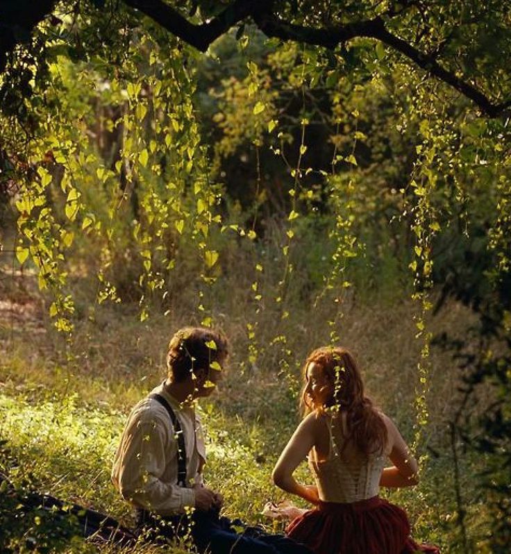 a man and woman sitting on the ground next to each other in tall grass with trees behind them