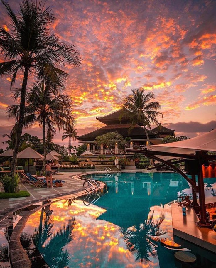 an outdoor swimming pool at sunset with palm trees and umbrellas in the foreground