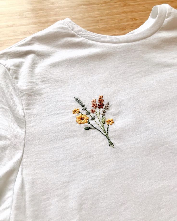 a white t - shirt with flowers embroidered on the front and back, sitting on a wooden surface