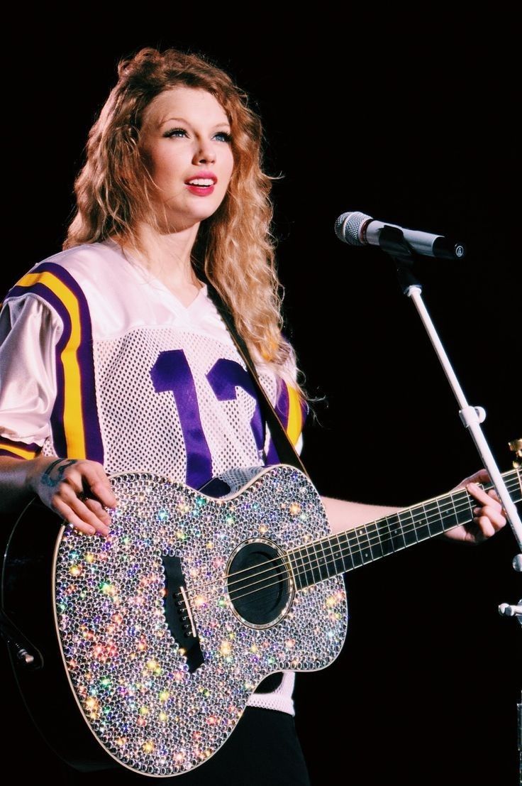 a woman holding a guitar while standing in front of a microphone and wearing a football jersey