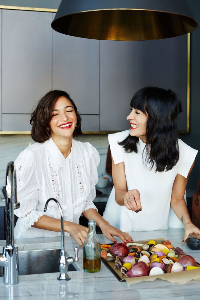 two women sitting at a kitchen counter cutting up vegetables on a cutting board and smiling