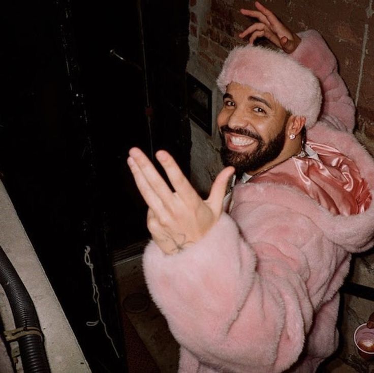 a man in a pink fur coat waving at the camera while standing next to a brick wall
