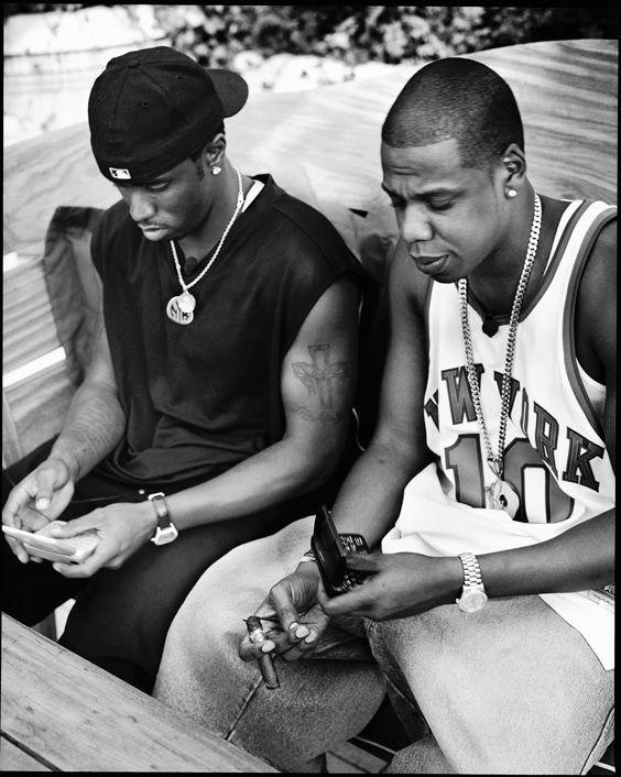 two young men sitting on a bench looking at their cell phones while one looks at his watch