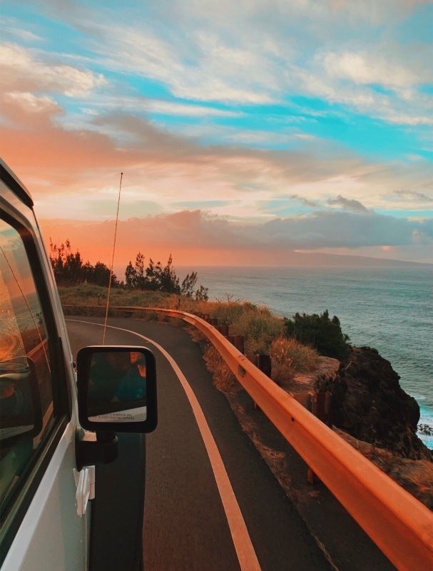 a car driving down the road next to the ocean at sunset or sunrise with clouds in the sky