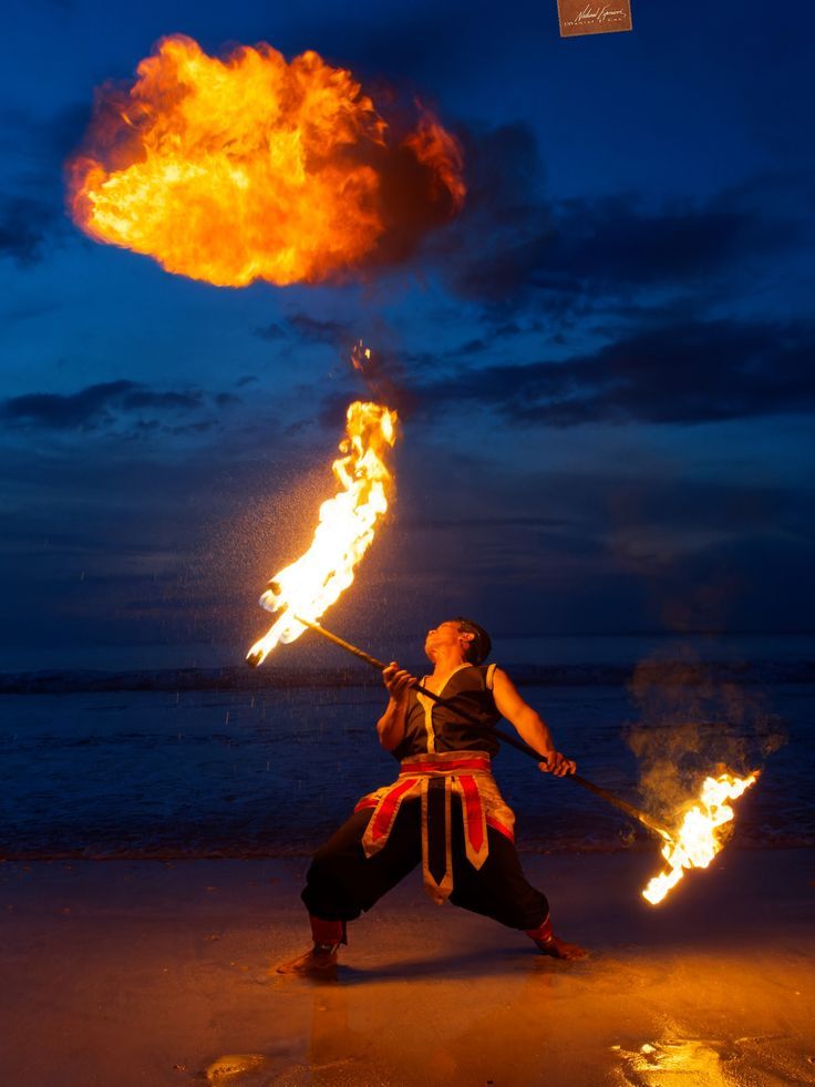 a man holding two torches in his hands on the beach at night with an orange flame behind him