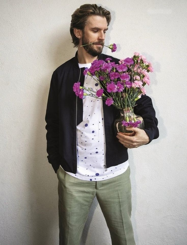 a man with flowers in his mouth standing against a wall holding a glass vase filled with pink flowers