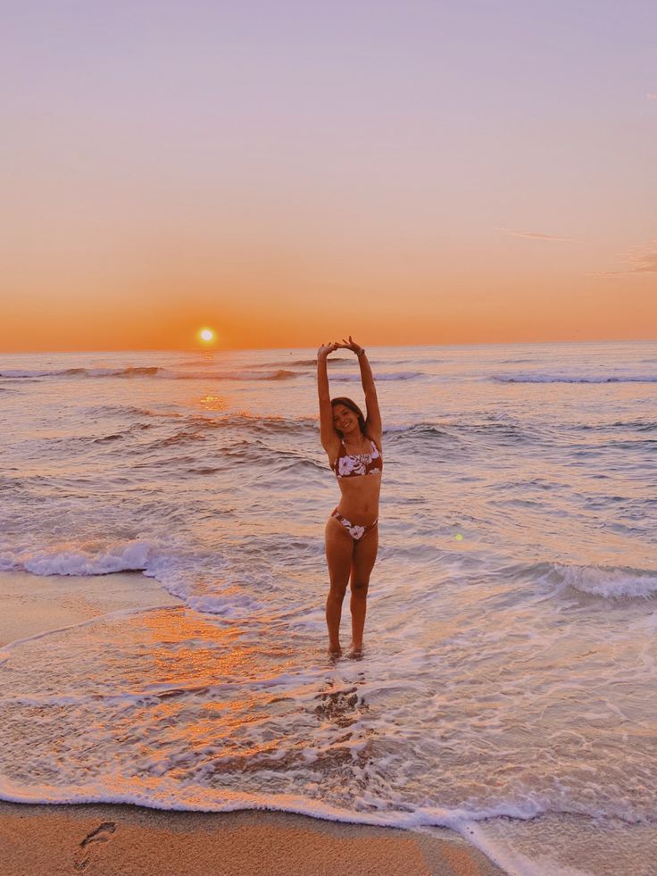 a woman standing in the ocean at sunset with her arms up and hands above her head