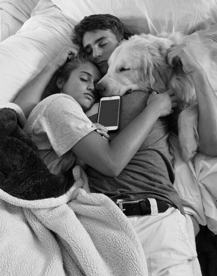 a man and woman laying in bed next to a dog with an electronic device on their lap