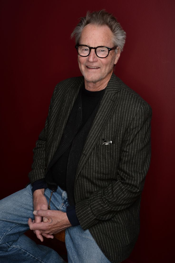 an older man wearing glasses sitting on a chair with his hands in his pockets and looking at the camera