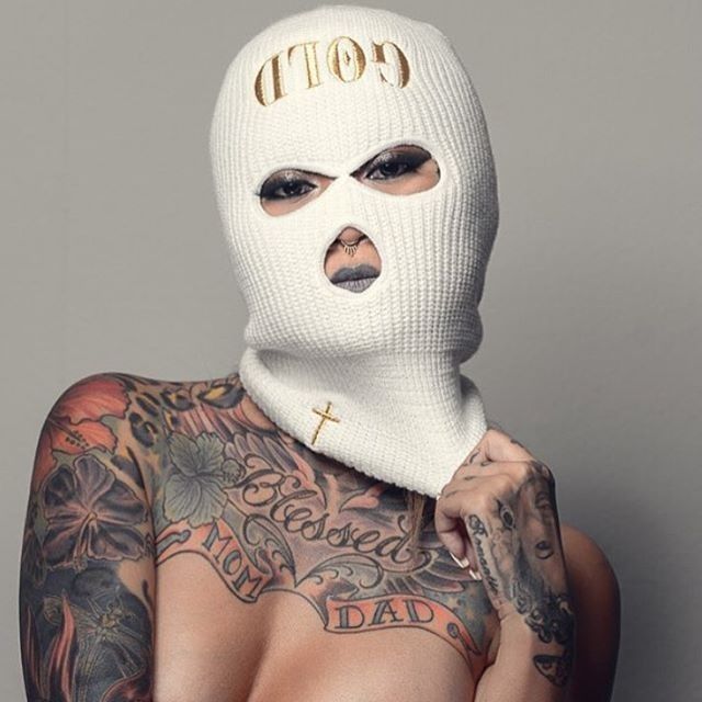 a tattooed man wearing a white mask with gold lettering on his face and upper half