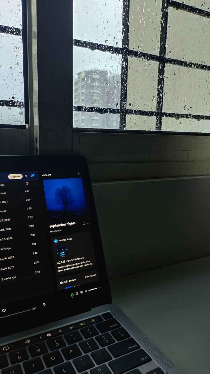 an open laptop computer sitting on top of a desk next to a window covered in rain