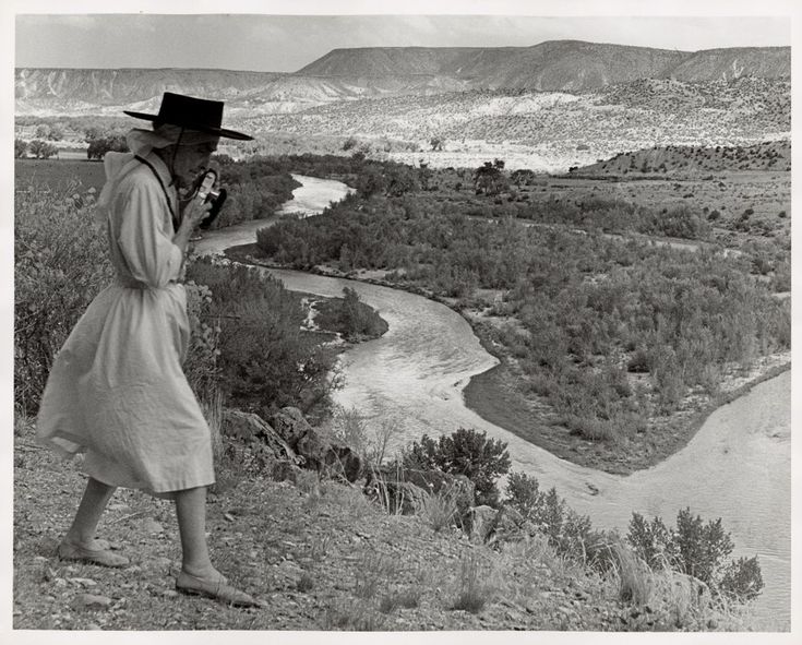 an old photo of a woman in a hat and trench coat standing on a hill