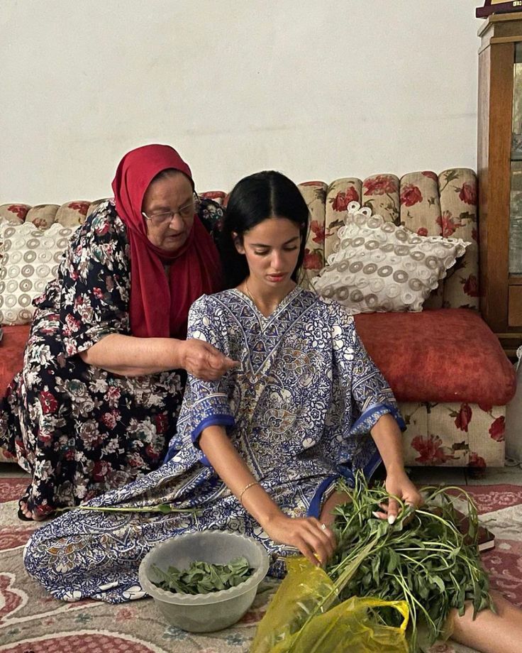 two women are sitting on the floor and one is cutting up some vegetables while the other looks at her phone