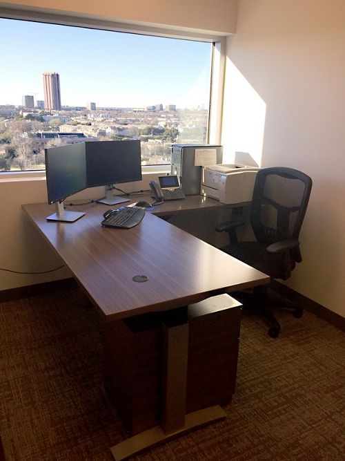 an office desk with two monitors and a keyboard in front of a large window overlooking the city